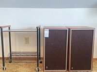 Front view showing pair of speakers with wooden frames and brown fabric covers alongside black record stand cart with wooden handles and metal wire rack lower shelf.