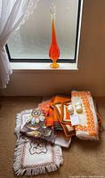 Tall orange graduated swung glass vase placed on a window ledge beside a set of folded vintage table linens and napkin rings.