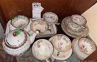 Lot of teapot, teacups, saucers, and serving plates arranged on a glass shelf showing the decorative patterns and condition including the teapot lid.