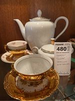 Porcelain teapot, sugar bowl, and gold floral teacups and saucers shown together on wooden surface.