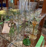 Shelf display with multiple clear wine and liquor glasses, some with hand-painted green and gold stems, varying sizes and shapes visible, including those with decorative touches and minor chips/fleabites.