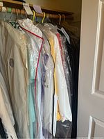Photo of formal dresses and jackets hanging on wooden rod inside a closet, some covered in clear protective garment bags, showing variety of colors and protected state.