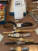 Seven women's wrist watches laid out on wood surface with some jewelry and boxes in background.