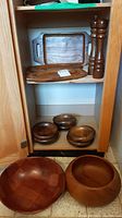 Two wooden serving trays and two tall spice shakers on upper shelf, three bowls on lower shelf inside cabinet, large checkered wooden bowl on floor outside cabinet