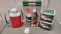 Photo shows two boxed Coleman stainless steel camping pot sets, a boxed Stanley insulated thermos set with two mugs, and a red insulated water jug with handle.