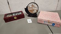 Three Asian collectable items arranged on table: rectangular bird inlay box, round black carved art, pink paper box