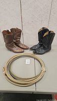 Two pairs of cowboy boots and a coiled lasso rope on a grey table against a light wall.