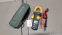 Greenlee CM-1550 clamp multimeter in yellow and black, accompanied by red and black test probes and a green zippered carry case with Greenlee logo.