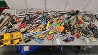 Wide view of lot showing various tools laid out on a table including hammers, staple guns, tape measures, drill bit sets, cutters, screwdrivers, pliers, levels, scissors, and small hardware.