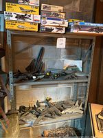 Photo showing assembled model airplanes on metal shelving, with various empty model kit boxes stacked above. Visible aircraft models include F-19 Stealth Fighter, Black Widow P-61, Lockheed SR-71A with D-21 Drone, P-40 Flying Tiger, F-15 Eagle, AH-64 Apache, B-25 Mitchell.