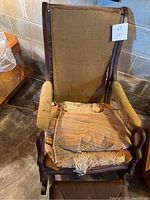 Antique rocker chair showing the back side, worn upholstery and seat cushion in poor condition with exposed padding.