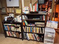 Two wooden bookshelves filled with a variety of books, framed certificates and awards on the wall, and miscellaneous office supplies including a network adapter and plastic drawer organizer.