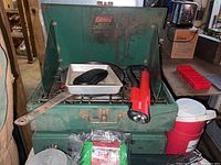 Green Coleman two-burner camping stove with metal drip tray, red flashlight, cooking spatula, and other camping supplies on and around the stove.