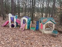 Wide shot of plastic children's playhouse with pink slide and attached structures in outdoor wooded area with leaf ground cover.