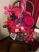 Red and purple hats, floral scarves, and accessories arranged on a wooden rocking chair, with red curtains in background and decorative items.
