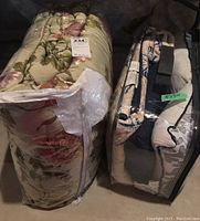 Two sets of bedding and cushions in plastic packaging placed on the floor. One is a floral patterned comforter set in light pastel colors by Croscill, the others are cushions by Madison Park in zippered bags.