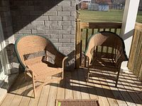 Two rattan patio chairs on a wooden deck by a brick wall and railing under partial sunlight