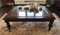 Full view of rectangular wood framed coffee table with clear glass top and woven cane panel below glass, turned legs and caster wheels, on patterned rug.