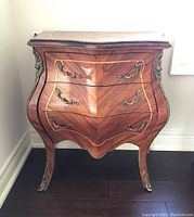 Front view of sculptured wooden end table with three drawers, ornate metal handles and trim, and inlaid veneer pattern.