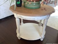 Side angle view of wooden end table showing distressed white painted base with fluted legs and circular lower shelf, and natural wood scalloped top with water marks, situated on dark floor.