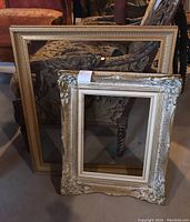 Two frames leaning against a patterned chair. Larger gold frame with ribbed design and smaller cream ornate frame with floral carving, both with glass.