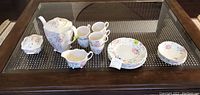 Full set arranged on glass table showing teapot, creamer, two cups, stack of plates and saucers