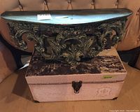 Overall view of wall mount sconce shelf sitting on top of Paris travel themed storage box on a cushion. Shelf carved with scrollwork, painted in distressed green.