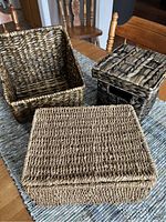 Three wicker baskets displayed on a table showing size and color differences. Small dark square basket with lid, medium open angle-shaped basket in dark tones, and larger rectangular light beige basket with lid.