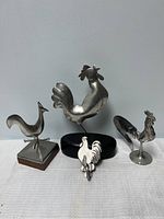 Four pewter rooster items: three standing figurines on bases of wood, glass, and oval shape, plus one flat rooster wall hook.