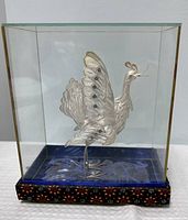 Front view of silver filigree bird sculpture inside glass case on blue fabric base with patterned lower trim