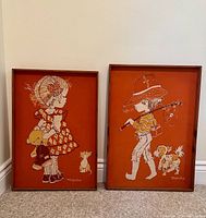 Pair of framed batik fabric artworks by Marsha on an orange background depicting a girl and a boy with pets, leaning against a wall.