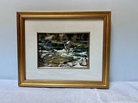 Photo of the framed painting showing a buck standing midstream in a rocky river, gold frame and cream matte visible.