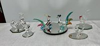 Front and angled view of six crystal rooster miniatures, including two colored Murano style pieces and four clear crystal pieces on mirror bases.