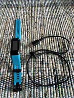 Photo showing blue Garmin fitness watch with rectangular screen and accompanying black charging cable on woven fabric surface.