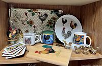 Wooden cutting board with rooster design, ceramic mugs with rooster prints, coasters with rooster designs, brass rooster napkin holders on a wooden shelf with rooster motif serving tray in background.