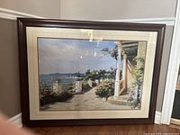 Full view of framed coastal landscape print featuring a walkway by the sea with plants, pillars, and distant water under a partly cloudy sky.