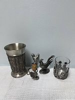German pewter tumbler, wine bottle stopper, two small pewter rooster figurines, and a pewter and glass candle holder with rooster motif all placed on a white cloth against a plain wall.