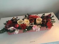 Pink stained wooden rectangular box holding Christmas-themed tabletop centerpiece with candles, artificial flowers, pinecones, berries, and ribbons.
