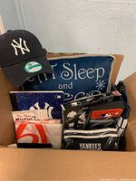 Box containing various New York Yankees merchandise items including a cap, a pillow with white text, packaged socks and tie, and notebooks.