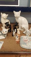 Various ceramic and china cat figurines, including large white cats, black bookend cats, and small colorful cats alongside a cat face pot holder