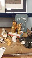 Photo of various ceramic and wooden cat figurines on a table including a black and white ceramic cat, a yellow striped ceramic cat, a solid white ceramic cat, and two larger wooden cat figurines painted as lion and tiger.