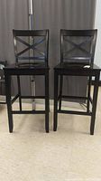 Front view of two black wooden bar height chairs with X-back design, showing scratches and wear.