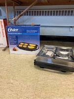 Oster folding electric grill in original box, displayed with parts of the grill visible beside the box on floor.