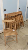 Set of 10 wooden folding chairs stacked against a wall with one chair unfolded in front.