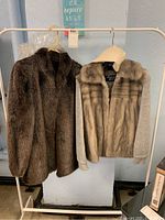 Photo shows two fur jackets hanging on a rack, one dark brown full fur and one mixed knit and fur jacket with light brown fur front and sleeves knit in cream color.