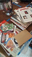 Photo showing a stack of Phillies magazines, newspapers, and assorted memorabilia spread on a table including card books and envelopes.
