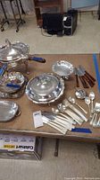 Full view of assorted silver-plated serving ware and cutlery including covered dishes, trays, ladles, and spoons laid out on table.