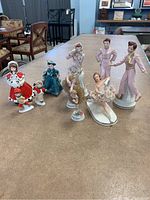 Ten skating figurines of varying styles and poses on a brown table in a room with chairs and shelving in background.