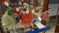 Four assorted skating figurines displayed on a glass shelf. Colorful ceramic sculptures depict female skaters in detailed period costumes.