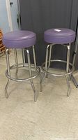 Pair of chrome stools with round purple faux leather seats showing rust and wear, standing on a tiled floor.